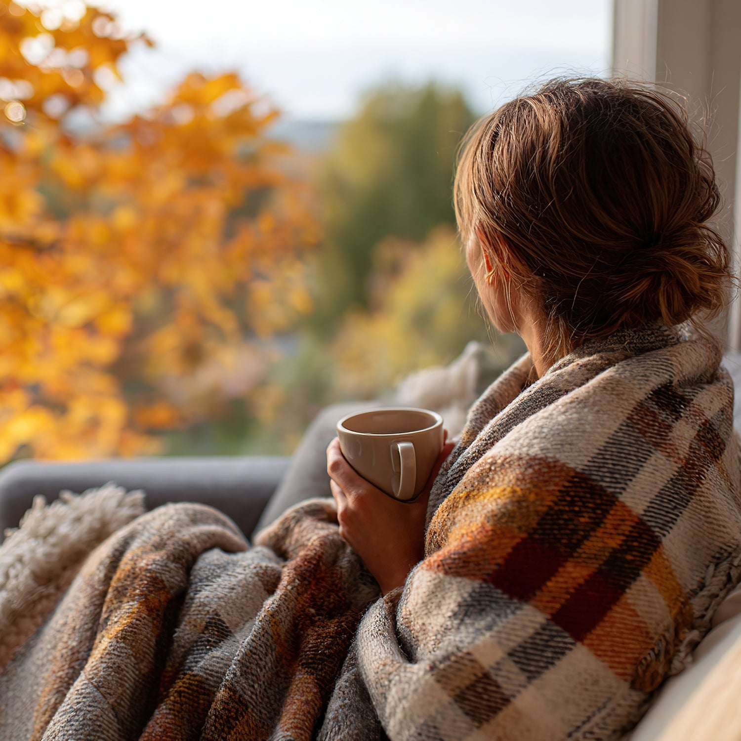 Cozy Fall - Woman wrapped in orange grey and white plaid fall blanket holding a coffee mug looking at orange fall foliage