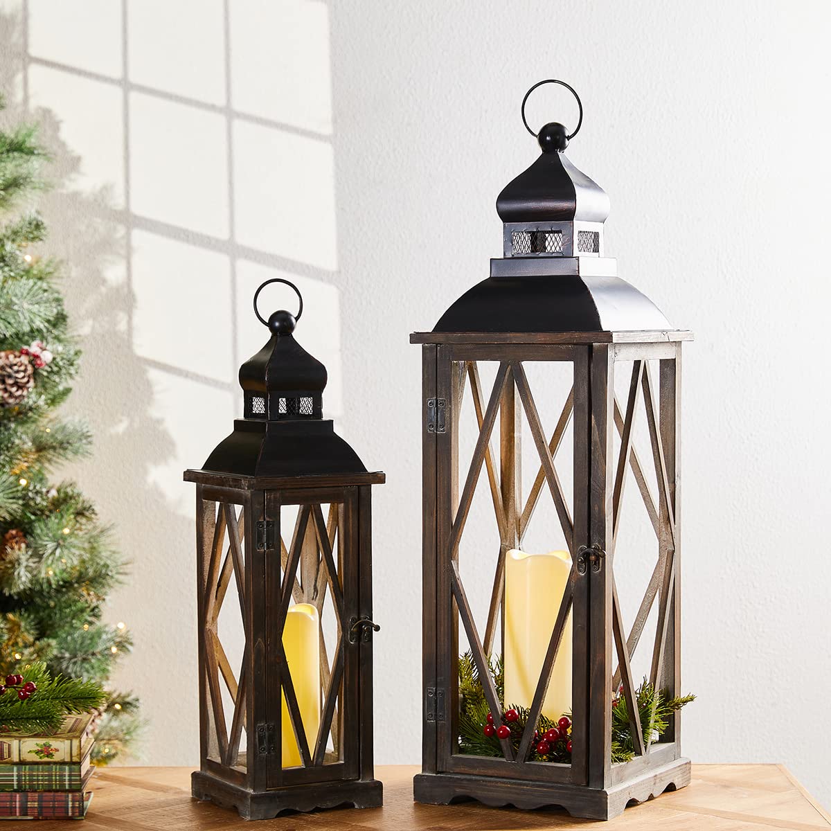 Two decorative lanterns with candles inside, one larger, on a surface with a Christmas tree in the background.