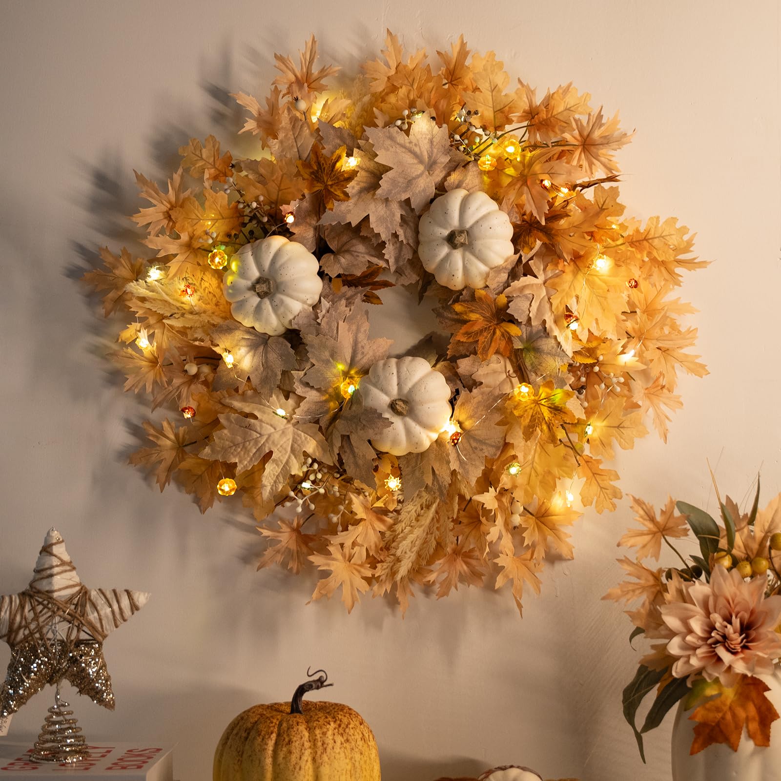 Decorative wreath with pumpkins and lights on a wall, with fall decorations on a table below.